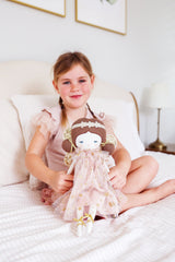 Girl sitting on white bed holding 40cm blush gold fairy doll with floral dress and wings.