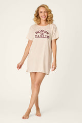 A smiling person with curly blonde hair wears the P.J. Salvage Saddle Up Howdy Darlin Nightshirt, a beige T-shirt dress featuring “HOWDY DARLIN” in maroon lettering, while standing barefoot against a plain, light background.