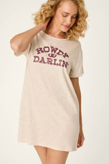 A woman with curly blonde hair wears the P.J. Salvage Saddle Up Howdy Darlin Nightshirt, a beige t-shirt dress featuring the phrase “HOWDY DARLIN” in red letters. She is smiling and looking down with one arm raised against a plain background.